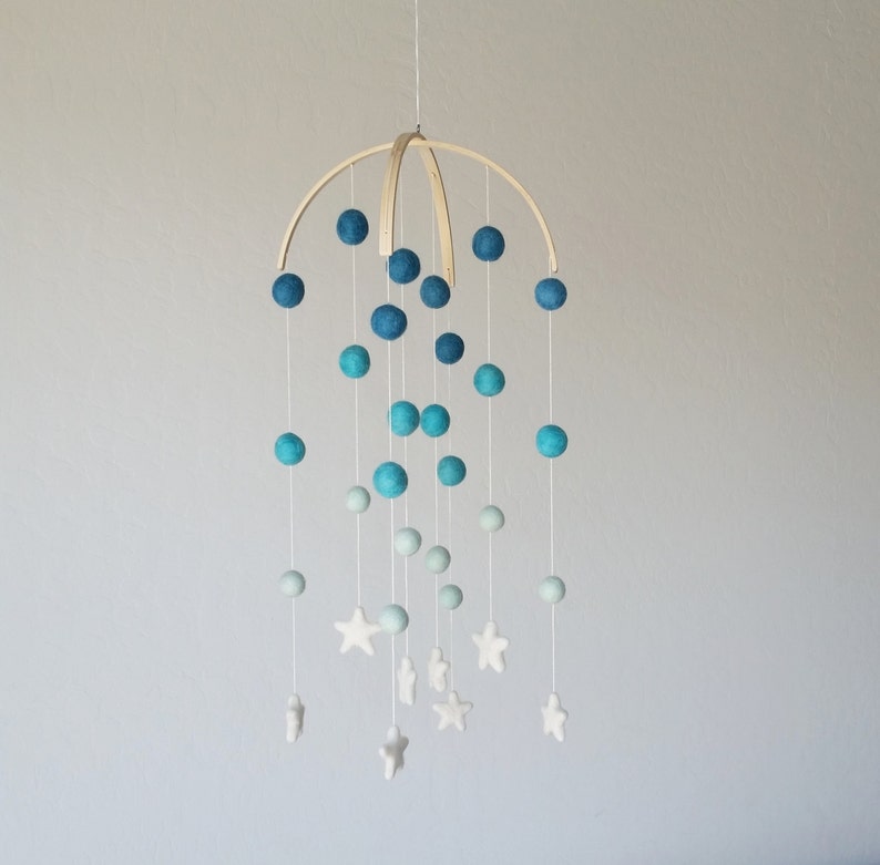 Op de afbeelding: Een houten babymobiel met een blauw-wit kleurenschema. De mobiel heeft viltballen in tinten blauw, overgaand in wit, en witte viltsterren.