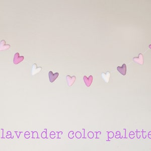 May include: A garland of felt hearts in shades of lavender, pink, and white. The hearts are strung on a white thread and hang in a gentle arc. The text "lavender color palette" is written below the garland.