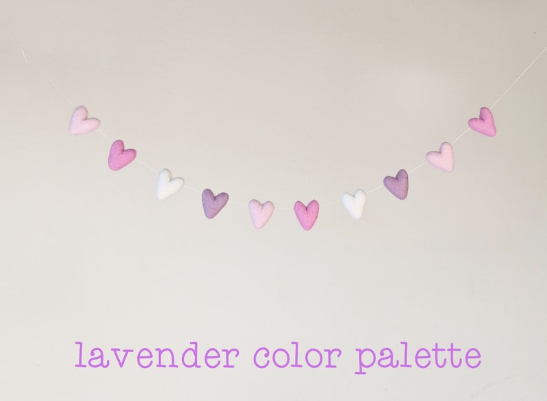 May include: A garland of felt hearts in shades of lavender, pink, and white. The hearts are strung on a white thread and hang in a gentle arc. The text "lavender color palette" is written below the garland.