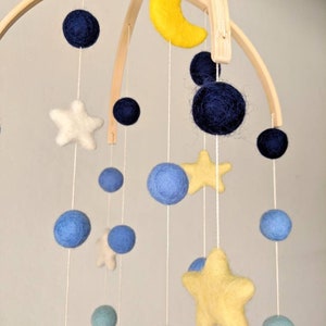 May include: A wooden baby mobile with felt stars, moons, and balls in blue, yellow, and white colors. The mobile is hanging from a wooden arch.