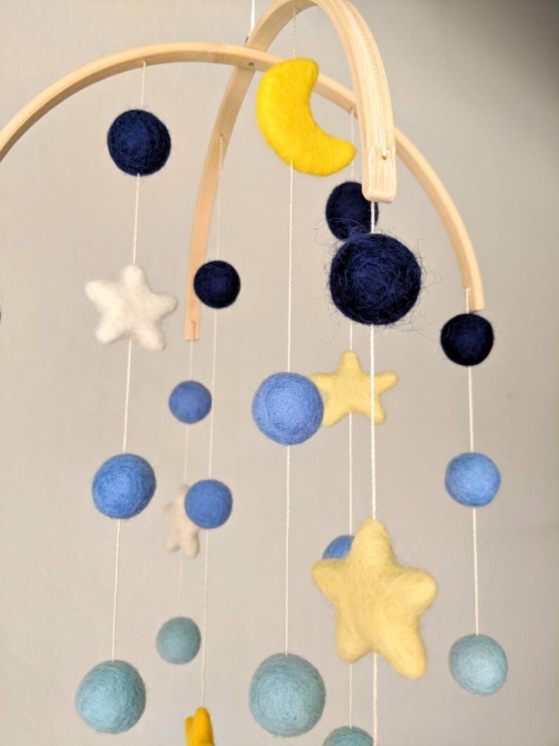 May include: A wooden baby mobile with felt stars, moons, and balls in blue, yellow, and white colors. The mobile is hanging from a wooden arch.