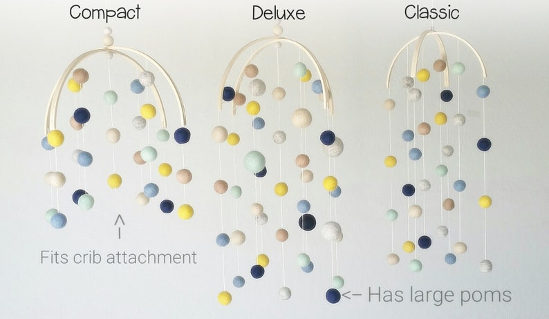 Op de afbeelding: Drie houten babymobielen met viltballen in verschillende kleuren, waaronder geel, blauw, groen en cr&egrave;me. De mobielen zijn gelabeld met "Compact", "Deluxe" en "Classic". De "Deluxe" mobiel heeft grotere viltballen dan de andere twee.