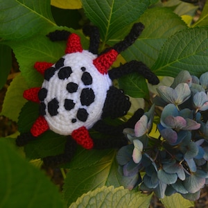May include: A handmade crochet spider toy with a white body, black spots, and red accents. The spider has black legs and is set against a backdrop of green leaves and blue hydrangea flowers. This unique toy is perfect for children.