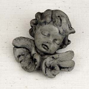 May include: A gray stone sculpture of a sleeping angel with wings. The angel has a cherubic face and is sculpted in a realistic style.