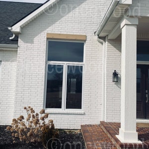May include: A white brick house with a black roof and a brown wooden trim above a large window. The house has a porch with a white column and a brick floor.
