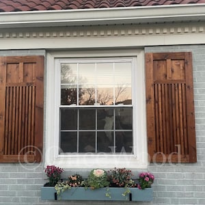 May include: Exterior view of a house with a window featuring wooden shutters and a window box. The shutters are a dark brown, and the window box is a light blue, filled with colorful flowers and greenery. The house has a gray brick exterior.