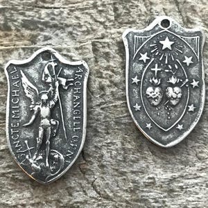 May include: Two silver-toned shield-shaped pendants. One features an angel with the text "SANCTE MICHAEL ARCHANGELI O.P.N." The other has a cross, stars, and hearts. Both have a small hole at the top for hanging.
