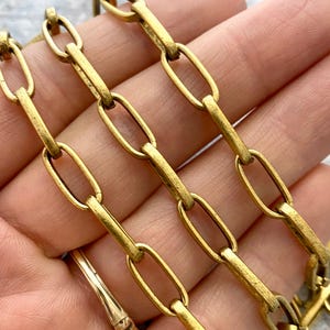 May include: Close-up of a gold-colored chain with elongated oval links. The chain is made of metal and has a vintage or antique appearance. The links are connected in a repeating pattern, creating a decorative element.