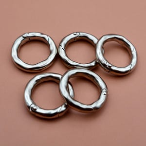 May include: Five silver-toned metal spring rings with a textured, organic, and slightly irregular circular shape. Each ring features a functional spring-loaded gate mechanism, allowing the circular clasp to open and close securely for attachment.
