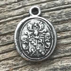Könnte beinhalten: Ein kleiner, silberfarbener Anhänger mit ovaler Form und detailliertem geprägtem Design. Das Design zeigt ein Wappen mit aufwendigen heraldischen Elementen, darunter ein Schild, ein Helm und dekorative Verzierungen. Der Anhänger hat oben eine kleine Schlaufe.