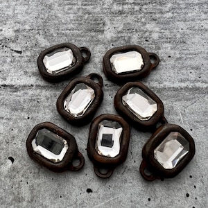May include: Seven antique-style rectangular charms with clear, faceted crystal centers. Each charm has a dark brown, metal frame with a small loop at the top for attaching to jewelry or other crafts. The crystals reflect light, creating a sparkling effect.