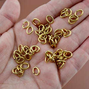 May include: A collection of small, circular gold-colored metal jump rings. The rings are scattered and some are linked together in small clusters. Each ring features a single cut point, allowing them to be opened and closed for jewelry making.