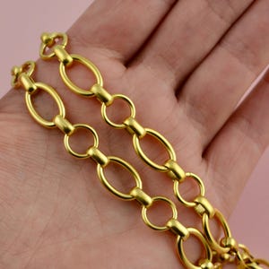 May include: Close-up of two gold-colored chain strands. Each strand features oval links connected by smaller, round links. The chains have a bright, shiny finish, suggesting they are made of metal. The background is a soft, blurred pink.