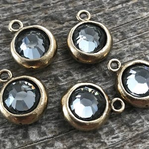 Peut inclure: Cinq breloques circulaires dotées d'un cristal gris foncé à facettes serti dans une monture en métal au ton laiton antique vieilli. Chaque breloque comprend un petit anneau de jonction sur le dessus, avec une finition métallique texturée.