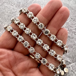 Puede incluir: Tres pulseras plateadas con cristales redondos y transparentes. Los cristales están engarzados en un bisel y unidos entre sí. Las pulseras se exhiben sobre una superficie gris.