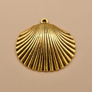 May include: A gold-toned metal pendant shaped like a scallop shell. The surface features a series of raised, radiating ridges that extend from the top center to the scalloped bottom edge. A small circular loop is attached at the top for hanging.