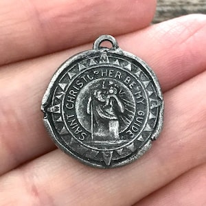 Op de afbeelding: Een ronde, zilverkleurige hanger met de tekst "SAINT CHRISTOPHER BE MY GUIDE" rond een centraal beeld van een figuur die een ander draagt. De hanger heeft een kleine lus aan de bovenkant om aan een ketting te bevestigen.