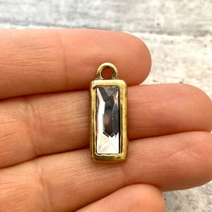 Puede incluir: Un dije rectangular dorado con un centro de cristal claro y rectangular. El dije tiene un pequeño lazo en la parte superior para sujetarlo a un collar o pulsera. El cristal refleja la luz, creando un efecto brillante.