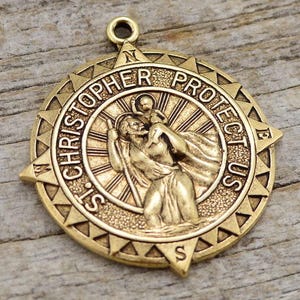Puede incluir: Medalla dorada de San Cristóbal con las palabras "ST. CHRISTOPHER PROTECT US" alrededor del borde. El centro representa a San Cristóbal llevando a un niño. La medalla tiene un lazo en la parte superior para colgar.