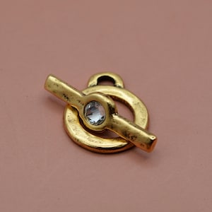 May include: A gold-toned toggle clasp featuring a circular ring and a T-bar component. The metal surface has a distressed, weathered texture. A single, clear, faceted circular crystal is embedded in the centre of the T-bar piece.