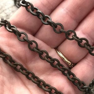 May include: Close-up of a bronze-coloured chain with oval links. The chain is held in a hand, with a gold ring visible. The chain is a decorative element, suitable for jewellery or craft projects.