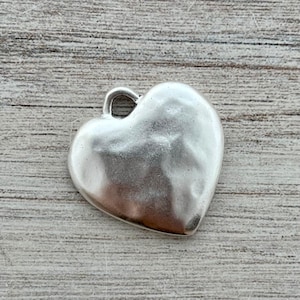 May include: A silver-colored heart-shaped charm with a textured surface and a small loop at the top. The charm is made of metal and is displayed on a light wood-grain surface.