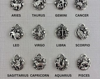 May include: A set of twelve circular silver-toned metal charms, each featuring a unique embossed zodiac sign symbol surrounded by stars. The charms are arranged in three rows of four, with the corresponding names Aries, Taurus, Gemini, Cancer, Leo, Virgo, Libra, Scorpio, Sagittarius, Capricorn, Aquarius, and Pisces printed below each piece.