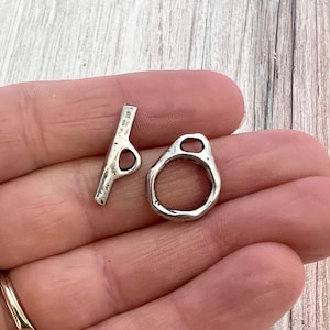 May include: A silver-toned toggle clasp set consisting of a T-bar component and a circular ring component. Both pieces feature a textured, uneven surface with a metallic finish, designed for use as a jewelry fastener or closure mechanism.