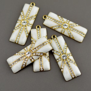 Puede incluir: Cinco colgantes rectangulares blancos y dorados con un diseño de estrella. Cada colgante presenta una pedrería transparente central rodeada de metal dorado y pedrería más pequeña. Los colgantes tienen un lazo en la parte superior para sujetarlos a una cadena u otra joyería.