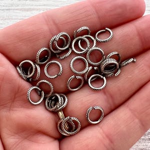 May include: A collection of small, silver-colored jump rings with a textured, rope-like design. The rings are open and ready for use in jewelry making or other crafts. The rings are held in a hand.