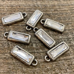 May include: Silver-toned rectangular charms with clear rectangular crystals. Each charm has a small loop at the top for attaching to jewellery. The charms are scattered on a wooden surface.