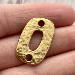 May include: A gold-toned metal connector charm with an oval shape and a textured, hammered surface. The piece features a large central oval cutout and two small circular holes at the top and bottom ends for attachment in jewellery making.