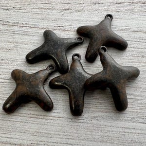 May include: Five dark brown, four-pointed cross-shaped metal charms. Each charm features a smooth, rounded surface with a small circular loop at the top for attachment. The metal has a weathered, matte finish with subtle variations in color tone.