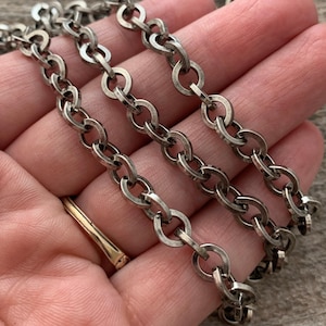 May include: Close-up of a silver-toned chain necklace with oval links. The chain is displayed in a hand, showcasing its texture and design. The necklace appears to be vintage or antique, with a slightly tarnished finish.