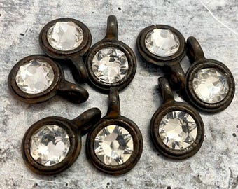 Könnte beinhalten: Eine Ansammlung von sieben runden Metallanhängern, die jeweils ein dunkles, verwittertes bronzefarbenes Finish und einen einzelnen, facettierten klaren Kristall in der Mitte aufweisen. Jeder Anhänger hat eine kleine Öse an der Oberseite.