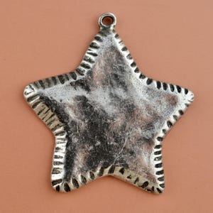 May include: A five-pointed star pendant with a distressed silver finish. The surface has a textured, uneven appearance with dark oxidation in the recessed areas. A small circular loop is attached to the top point for hanging or attachment.