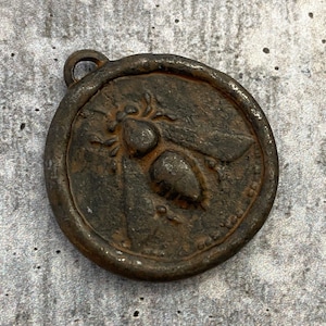 May include: A circular, dark brown, weathered metal pendant featuring a raised, embossed bee design in the center. The pendant has a textured, rustic finish and includes a small, integrated loop at the top for attachment to a chain or cord.