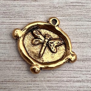 May include: A gold-colored dragonfly charm with a detailed design. The charm is circular with a raised border and a small loop at the top for attaching to a necklace or bracelet. The dragonfly is centered within the circle.