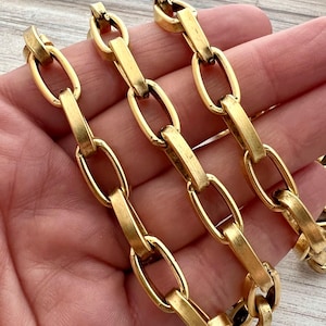 Puede incluir: Primer plano de un collar de cadena de color dorado. La cadena presenta eslabones ovalados alargados conectados por barras rectangulares. Los eslabones y las barras tienen un acabado pulido, que refleja la luz. El collar se sostiene en una mano, mostrando el diseño de la cadena.