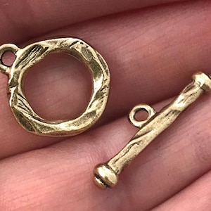 May include: A set of two brass-colored jewelry clasps. One is a textured ring with a loop, and the other is a bar with a loop and a rounded end. The clasps are designed for jewelry making.