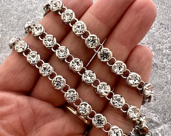 Könnte beinhalten: Ein silberfarbenes Metallkettenarmband mit einer durchgehenden Reihe runder, klarer, facettierter Strasssteine, die in einzelnen kreisförmigen Fassungen sitzen. Das Armband besteht aus verbundenen Gliedern, die ein flexibles Design bilden.
