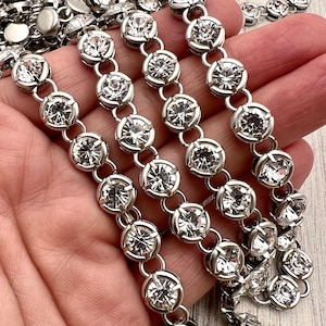 May include: Close-up of several strands of silver-toned rhinestone chain. Each chain link features a large, clear, round rhinestone. The chains are held in a hand, with the links arranged in parallel rows. The rhinestones sparkle.