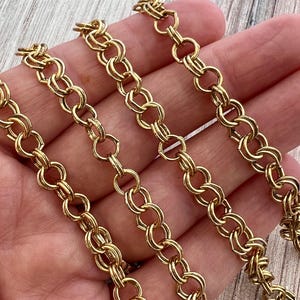 May include: Close-up of several strands of gold-colored chain. The chain links are a rounded, double-loop design. The chains are arranged diagonally across the frame, creating a pattern. The background is a light wood grain.