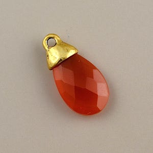 May include: A faceted, teardrop-shaped pendant with a reddish-orange gemstone. The top of the pendant is capped with a gold-colored metal loop for hanging. The gemstone has a polished, reflective surface.