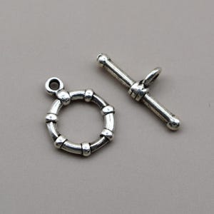 May include: A silver-toned toggle clasp set consisting of a circular ring with a segmented, beaded texture and a matching T-bar component. Both pieces feature a metallic finish with darkened recessed areas to highlight the structural details.