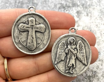 May include: Two circular silver-toned metal pendants featuring raised relief designs. One pendant displays a central cross symbol, while the other depicts a standing angel with wings. Each piece includes a small loop at the top for attachment.