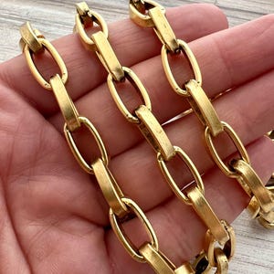 May include: Close-up of a gold-colored chain necklace. The chain features oval links connected by rectangular bars. The links and bars have a polished finish, reflecting light. The jewelry is likely vintage or antique.
