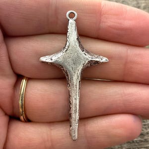 May include: A silver-colored metal cross pendant with a textured surface. The cross has four arms extending from a central square, with a loop at the top for hanging. The pendant is held in a hand.