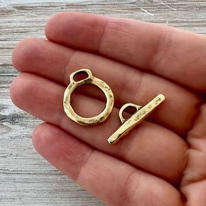 May include: Two brass toggle clasps. One is a ring with a small loop, and the other is a bar with a loop. The clasps have a textured surface and a warm, golden color. They are likely used for jewelry making or crafts.