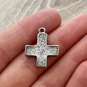 Puede incluir: Colgante de cruz plateada con superficie texturizada. La cruz tiene un lazo en la parte superior para sujetar a una cadena o cordón. El diseño es sencillo y clásico, adecuado para la fabricación de joyas o manualidades religiosas.